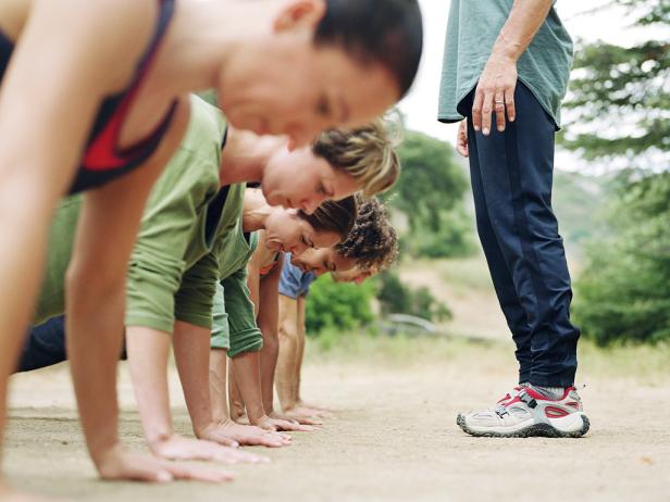  'Man standing over group of people doing pushups, side view'