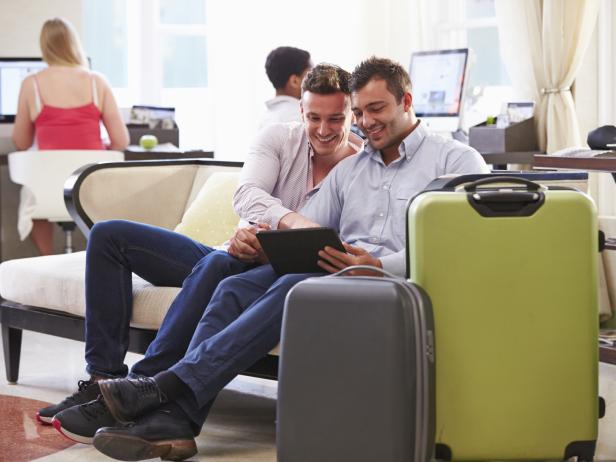Male Couple Sitting In Hotel Lobby Looking At Digital Tablet Smiling Male Couple Sitting In Hotel Lobby Looking At Digital Tablet Smiling