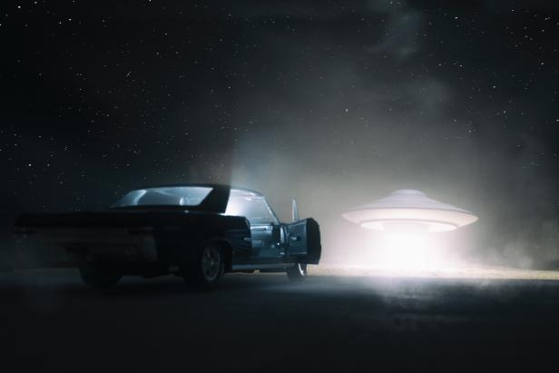 A UFO hovers over a desert, and an old car is parked in the foreground. 