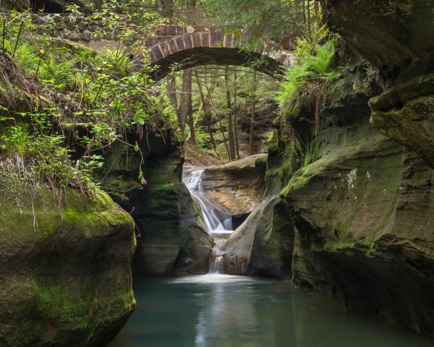 Peaceful landscape scene of waterfall gently falling in small gorge into pond with old brick bridge above surrounded by mossy boulders and spring green foliage