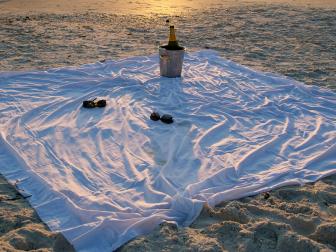 A big white sheet is laid out on the beach and there is an ice bucket and bottle of champagne with two pairs of sunglasses at sunset.