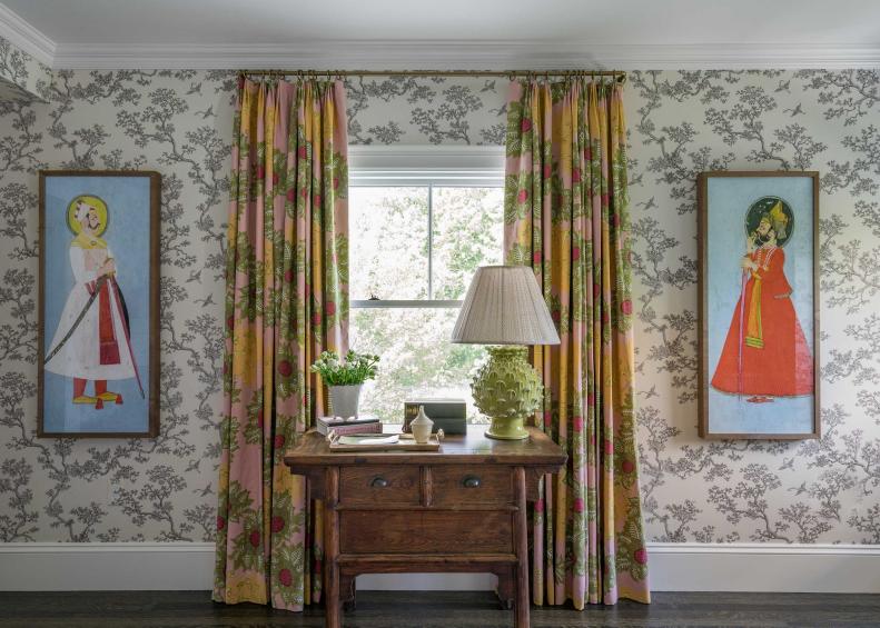 An antique desk sits before a window with floral drapes.