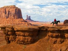 man rides horse along cliff in monument valley park with red rocks and caverns during the day