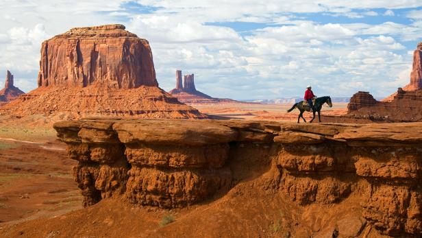 man rides horse along cliff in monument valley park with red rocks and caverns during the day