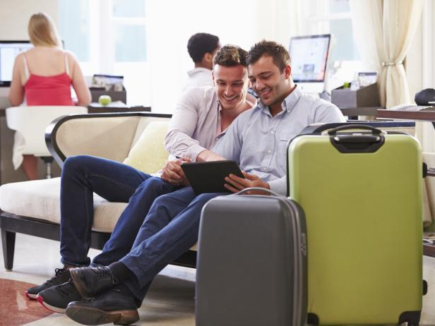 Male Couple Sitting In Hotel Lobby Looking At Digital Tablet Smiling