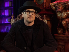 Zak Bagans is seated in an ornate chair wearing a black bowler hat, black jacket, and black t-shirt.