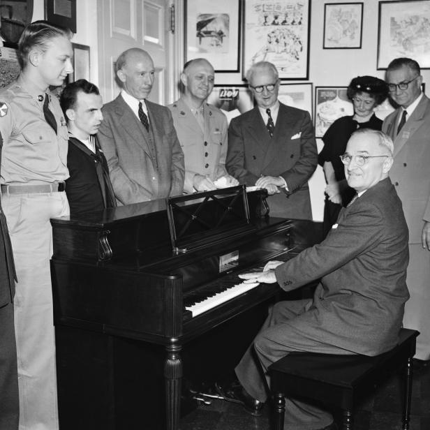 Black and white photo of President Truman seated at a piano surrounded by a group of seven men and one woman.