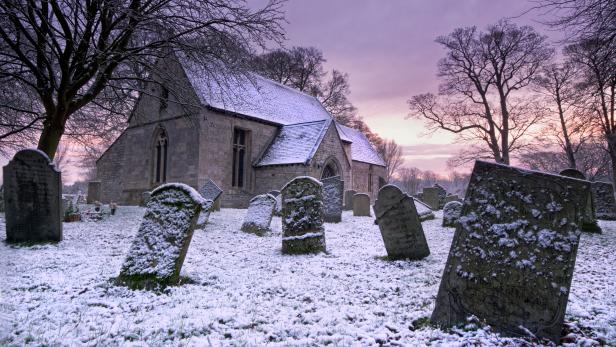 The colorful tones of a winter's sunrise can be seen across the sky in this English winter scene.Snow covers the ground and the gravestones of this Lincolnshire church at Little Ponton,UK. The church is seen with a snow covered roof. Trees add to the framing of this editorial image.
