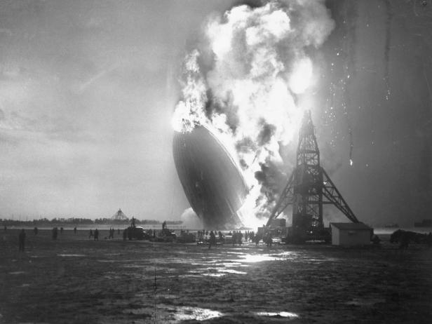 Members of the landing crew are shown fleeing from beneath the dirigible Hindenberg as the flaming craft, a greasy pall of smoke marking its last train, falls to the Lakehurst Landing Field.