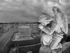 A praying statue kneels on top of a tomb in New Orleans under moody skies.  High point of view to contain the cemetery landscape in the background.