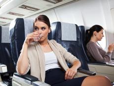 Young woman sitting on the airplane and drinking water, while woman in the background using a smart phone.