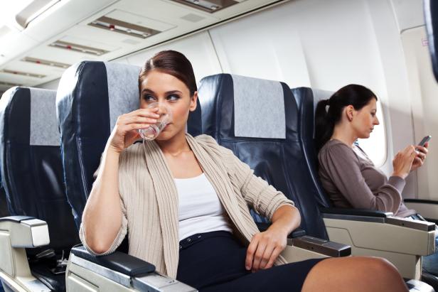 Young woman sitting on the airplane and drinking water, while woman in the background using a smart phone.