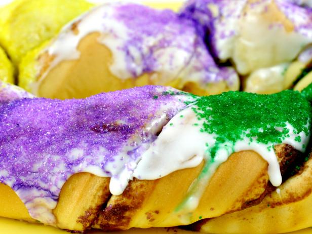"A close-up of a King Cake on a yellow background. A King Cake is a festive and traditional Mardi Gras treat more specific to New Orleans, LA. It tastes like huge cinnamon roll (you cut it in pieces just like other cakes) dripping with LOTS of icing and colored sugar on top (in green, purple and yellow -- the official Mardi Gras colors). YUM!"