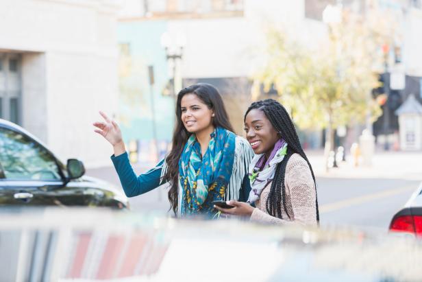 Two multi-ethnic young women in their 20s standing on a city street waiting for a car to pick them up. The African American woman is holding a mobile phone and her Hispanic friend is waving, trying to get the attention of their crowdsourced taxi.