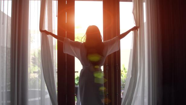 Young woman in bathrobe open curtains and stretch standing near the window at home. 4K