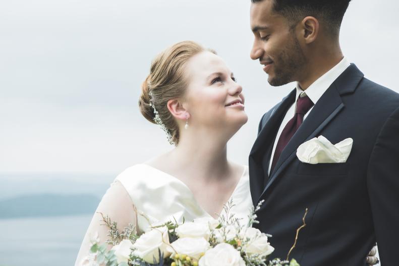 In this closeup, a beautiful bride stands outdoors with her groom and smiles up at him.  He looks down and smiles at her.  There is a scenic background.