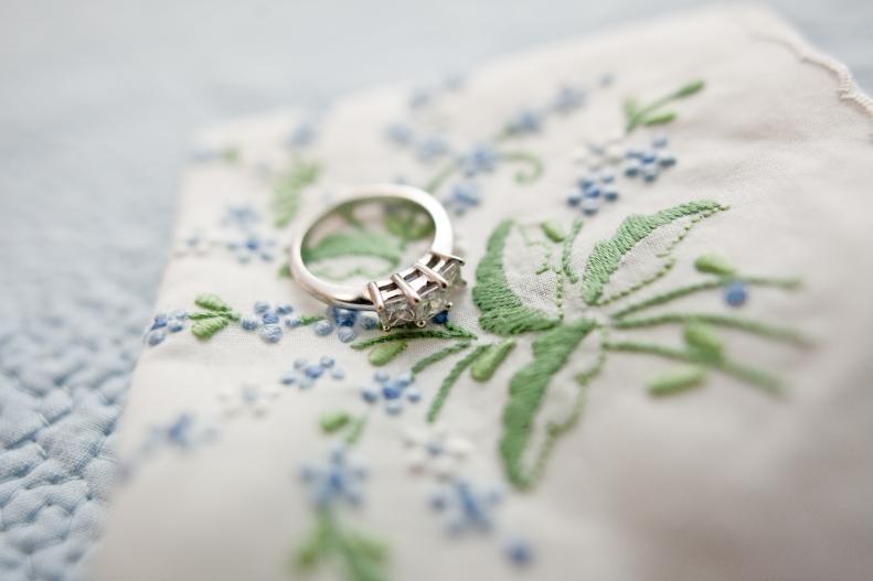 "A white gold, or platinum, engagement/wedding ring on a handkerchief.  The handkerchief has a small blue flower with green leaf embroidered design.  The handkerchief is resting on a lovely blue texture with a flower pattern."